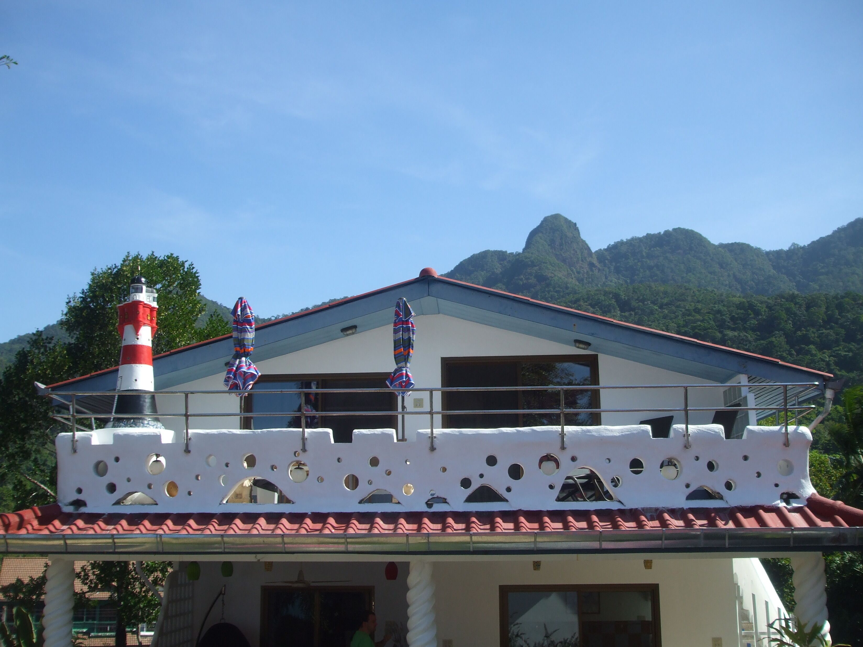 A building with a red and white lighthouse, a balcony with patterned umbrellas, and a mountainous backdrop.