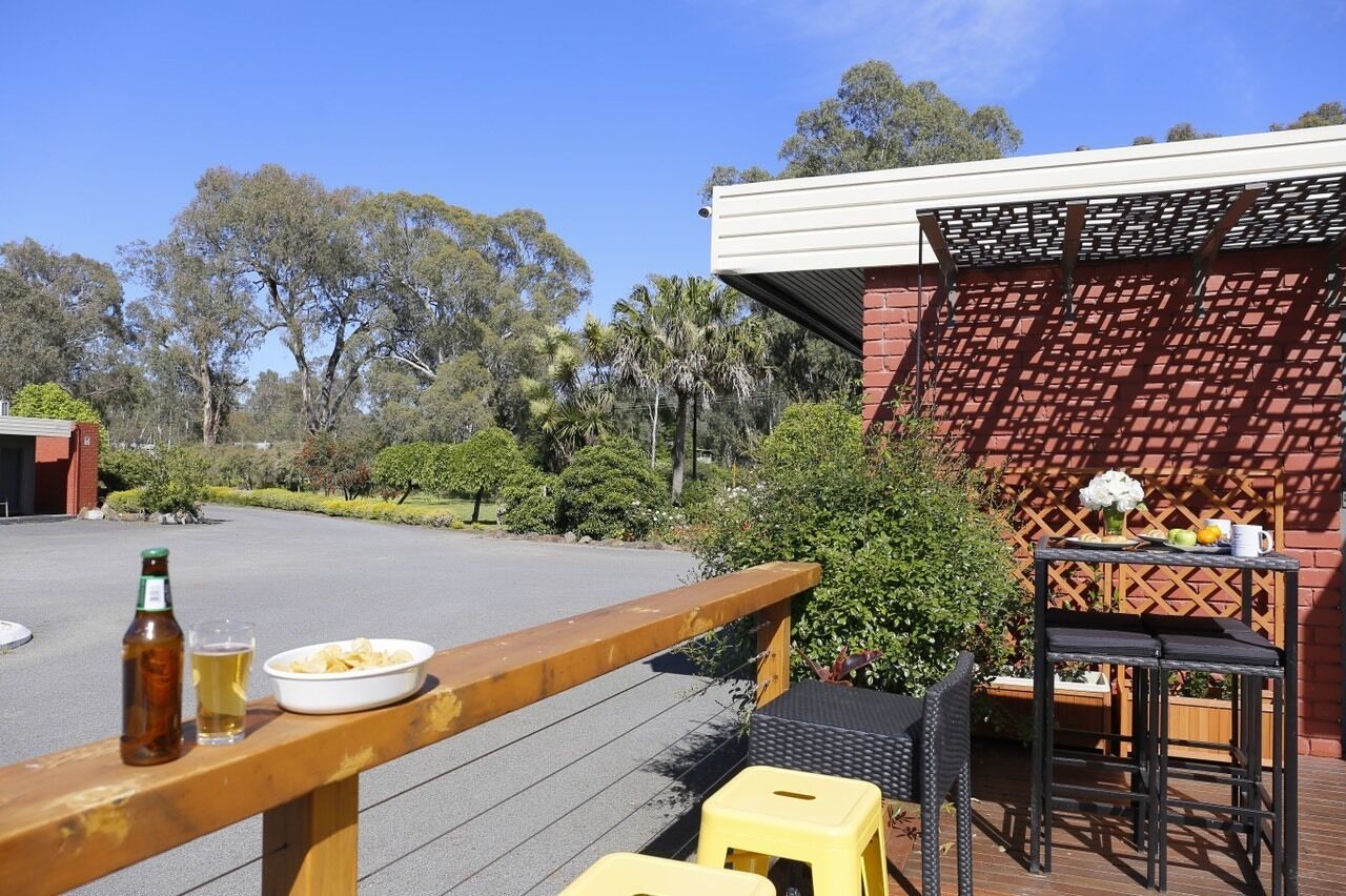 An outdoor seating area with a wooden deck, a table with a bottle and bowl of chips, and a small table with a vase and mug.