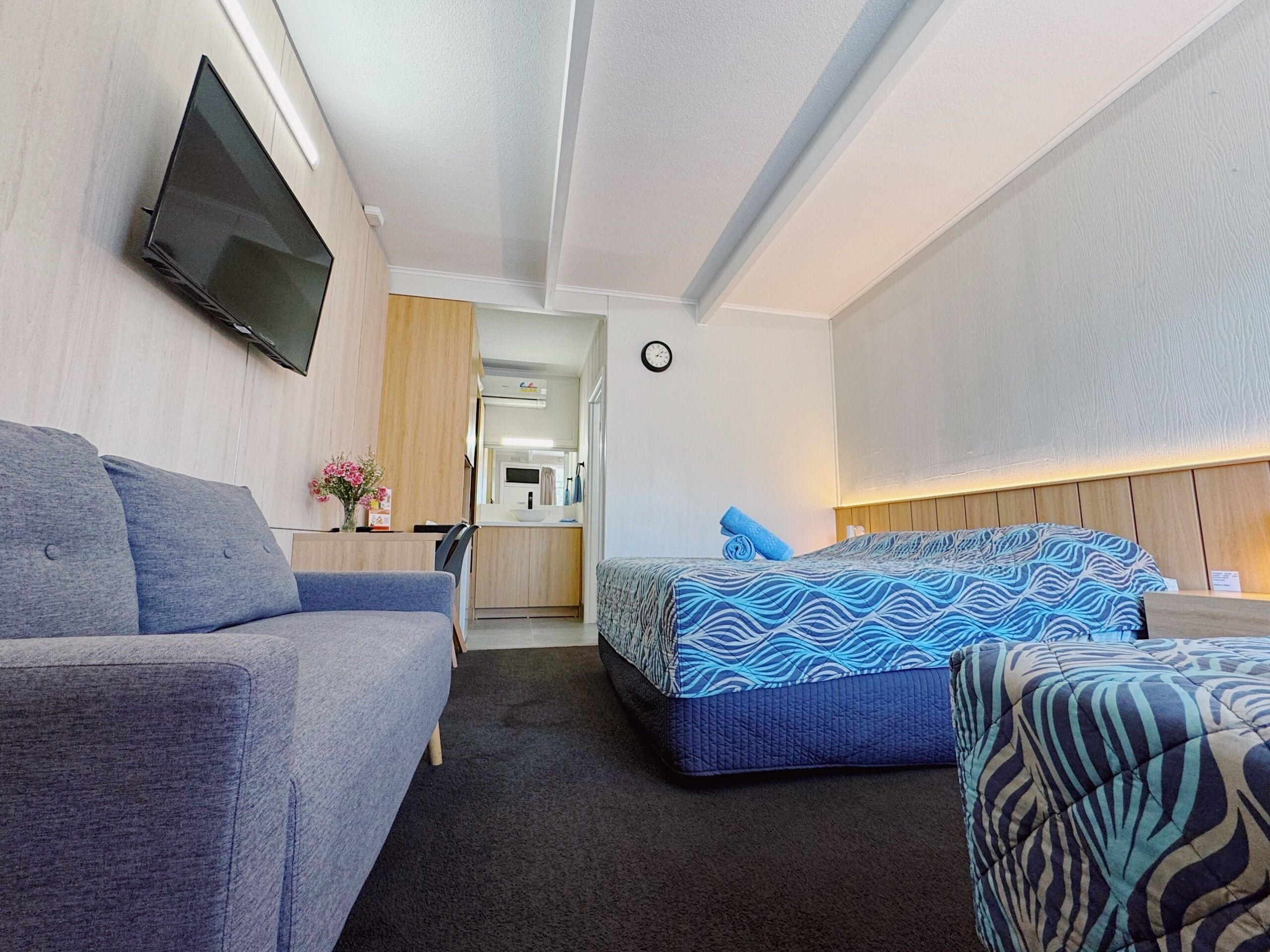 Habitación compacta con sofá, cama con ropa de cama estampada, televisor de pantalla plana montado en la pared y una pequeña kitchenette al fondo.