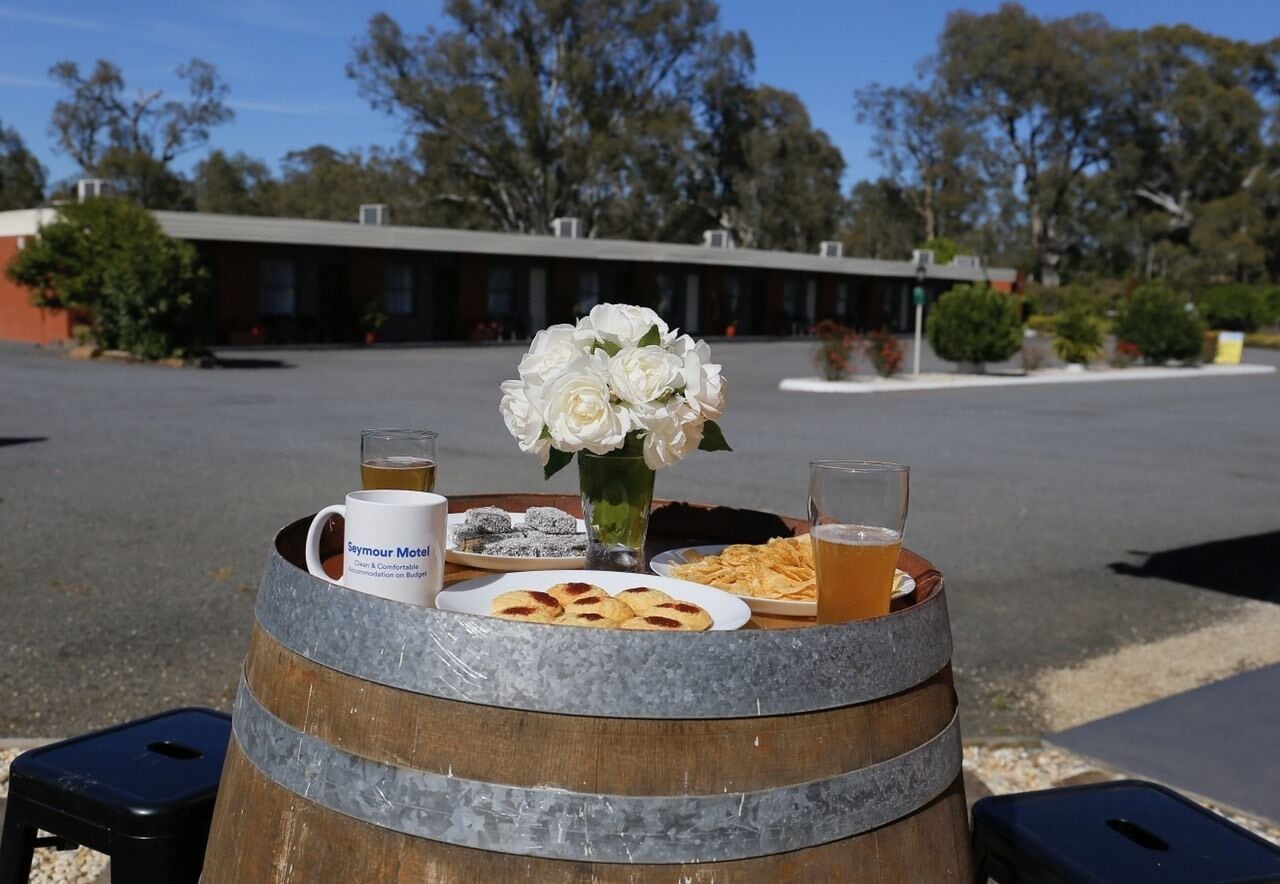 A round wooden barrel with a glass of beer, a mug, a plate of cookies, and a vase of white flowers on a sunny day.