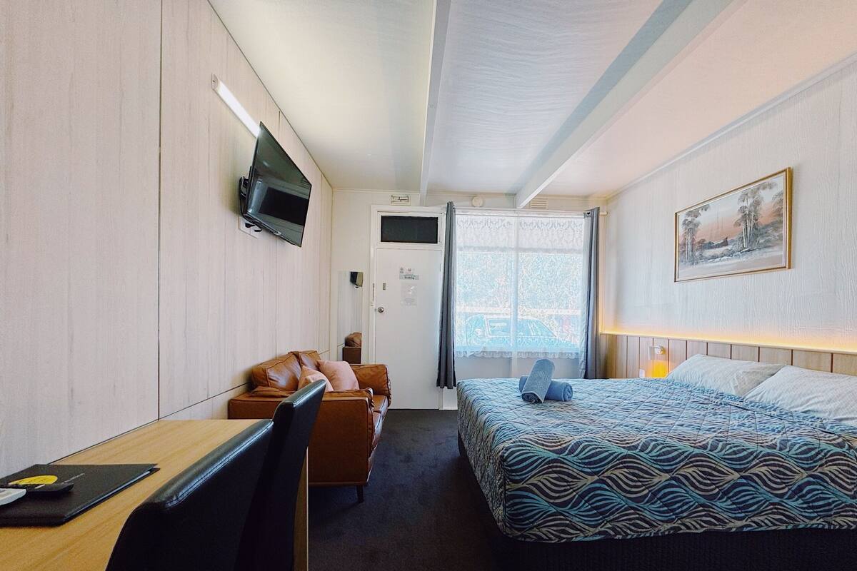 A hotel room with a bed, a desk, a chair, a TV mounted on the wall, and a window with curtains.