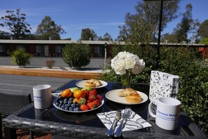 A table with breakfast items, a vase of white flowers, and a menu on a sunny outdoor patio. - Seymour Motel (Seymour)