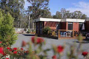 Un pequeño edificio de dos plantas con fachada de ladrillo y techo plano. Hay áreas de estar al aire libre con mesas y sillas, y un coche estacionado adelante.