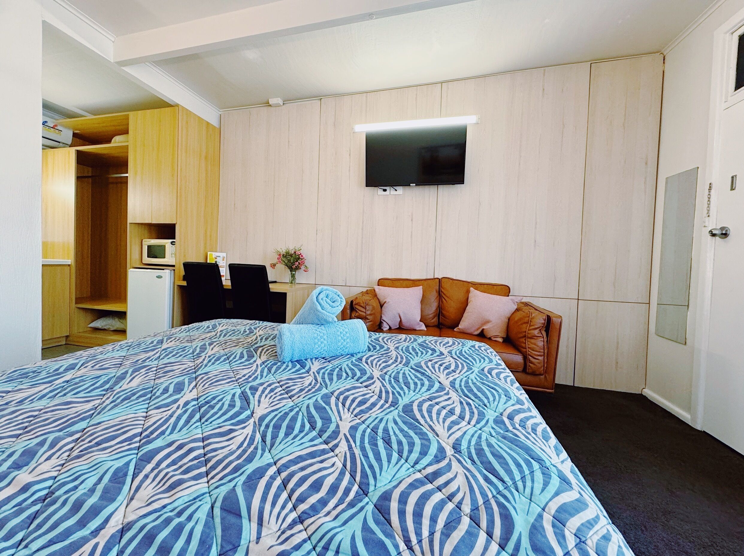 A hotel room with a bed, a TV mounted on the wall, a small kitchenette, and a seating area with a sofa.