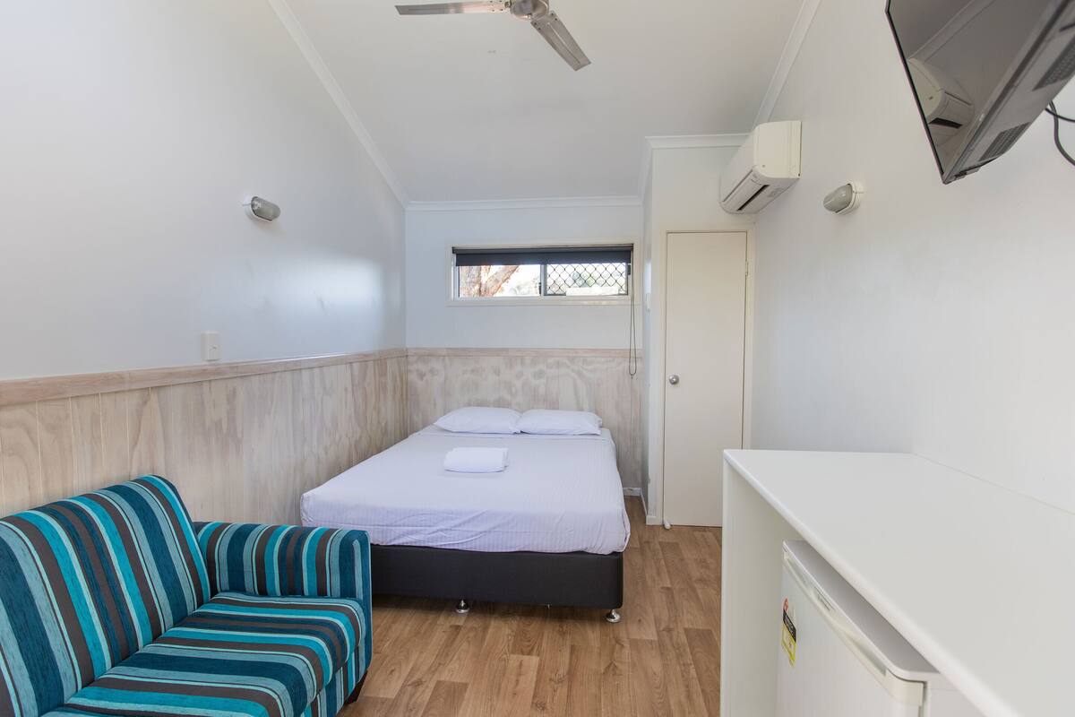 A bedroom with a bed, a striped sofa, a desk, a ceiling fan, a window, and an air conditioning unit.