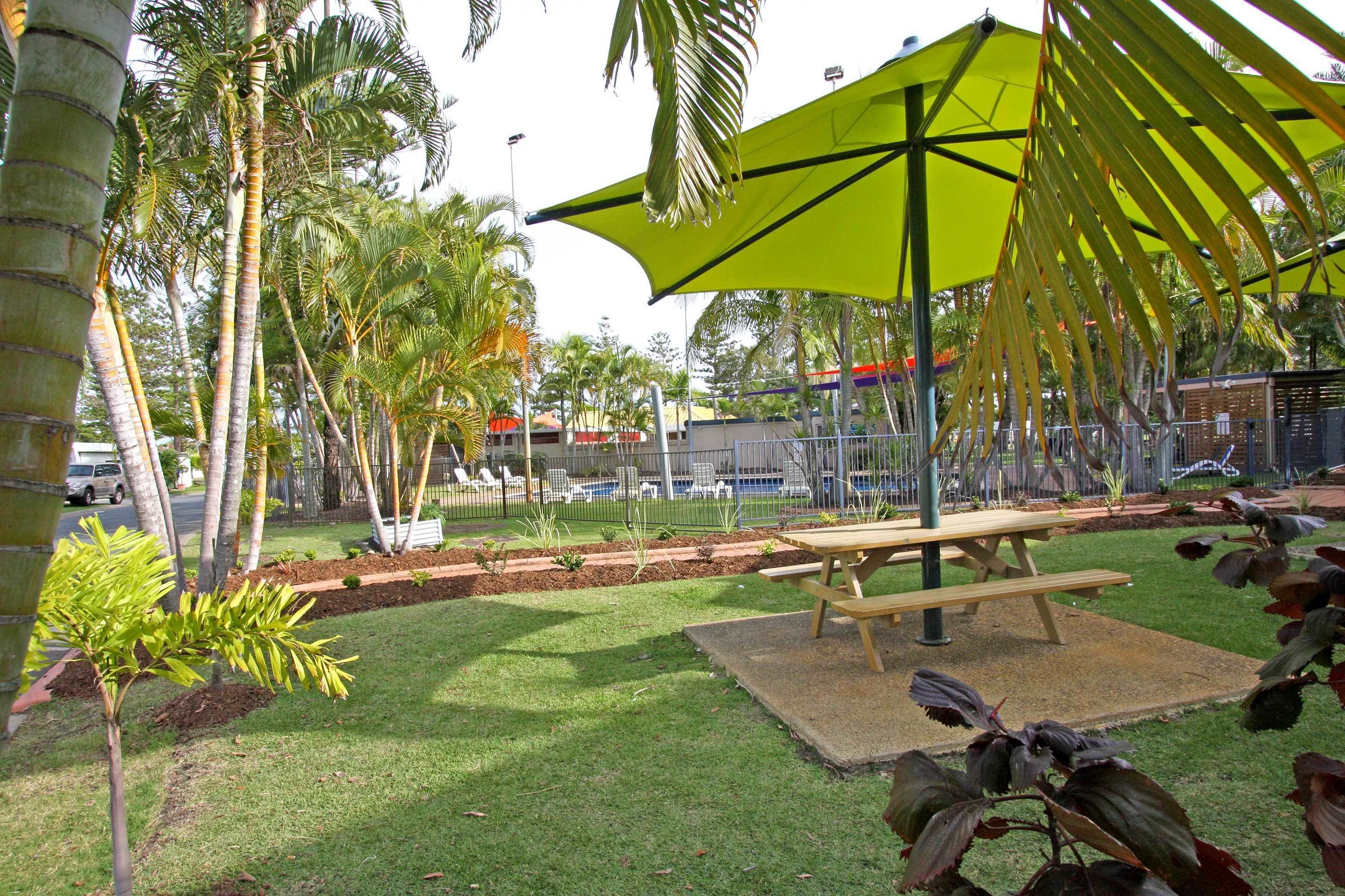 A park area with a picnic table, a large green umbrella, and palm trees.