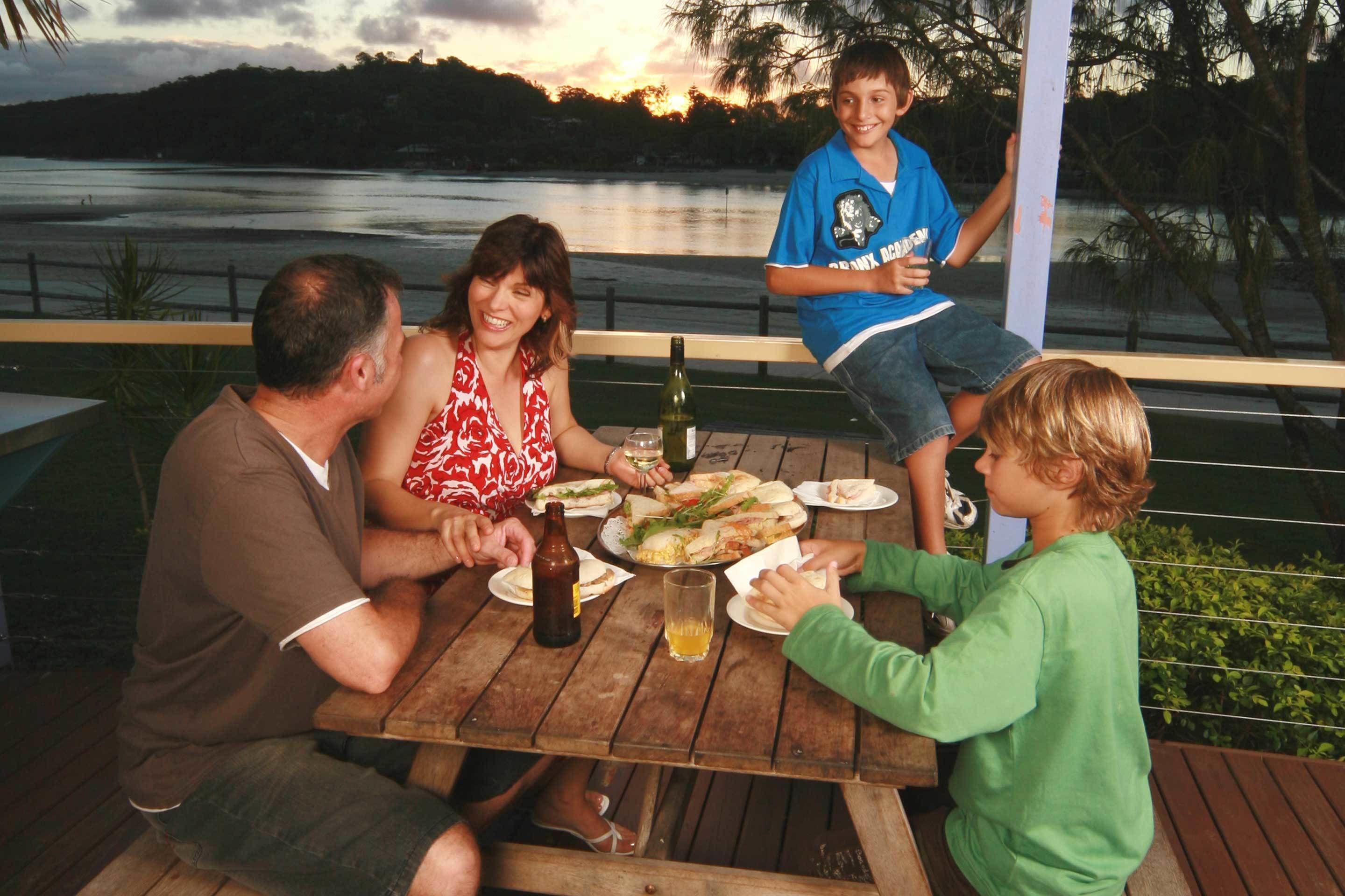 A group of people seated at a wooden table outdoors with a view of a body of water and trees in the background.