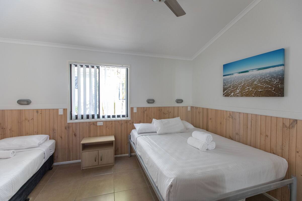 A bedroom with two beds, a window with blinds, a wooden wall panel, a painting, and a small wooden cabinet.
