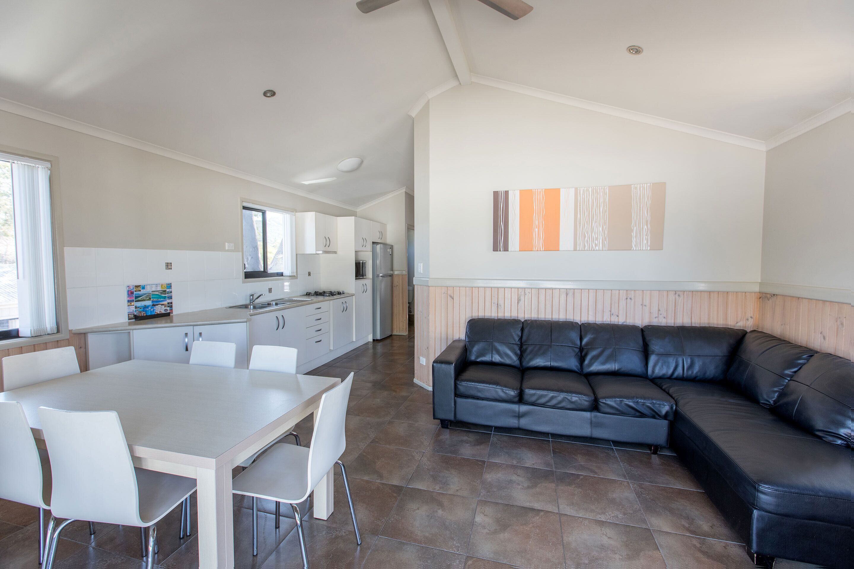 A spacious living area with a large black leather sofa, a dining table with chairs, and a kitchen in the background.