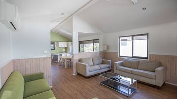 Ein großzügiger Wohnbereich mit einem grünen Sofa, beigen Couches und einem Glastisch. Ein Deckenventilator und große Fenster sorgen für natürliches Licht.