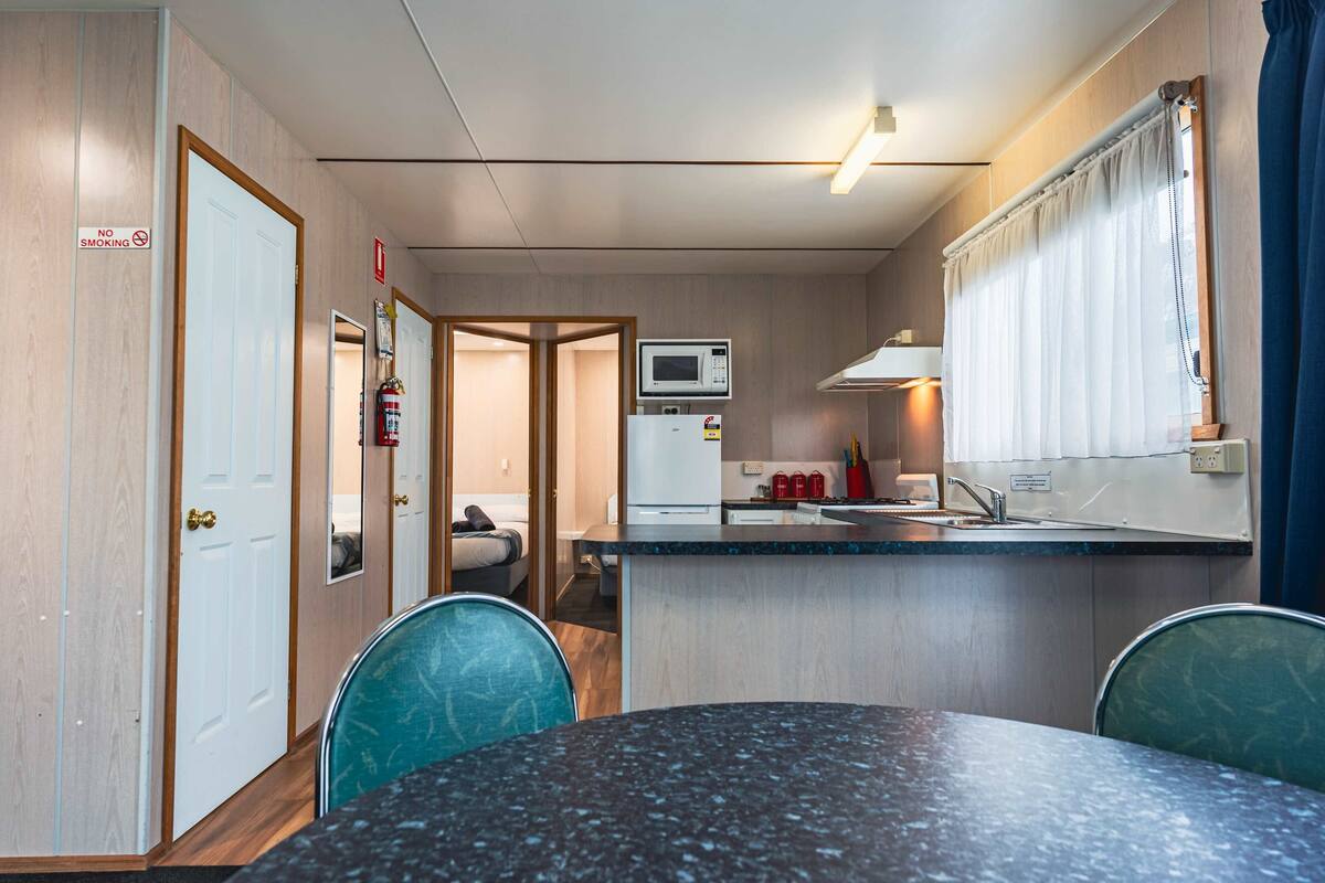 Una kitchenette compacta con microondas, pileta y heladera pequeña. Hay un comedor con mesa y sillas, y una puerta con un cartel de 'No Smoking'.