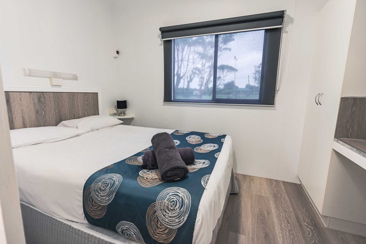 Una cama bien hecha con una colcha estampada y toallas enrolladas, una mesita de noche con lámpara, y una ventana con vista a árboles.