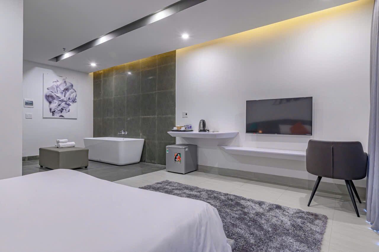 A modern bedroom with a large bed, a small bedside table, a gray ottoman, a gray rug, a large mirror, a wall-mounted TV, and a bathroom with a bathtub and sink.