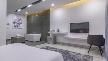 A modern bedroom with a large bed, a small bedside table, a gray ottoman, a gray rug, a large mirror, a wall-mounted TV, and a bathroom with a bathtub and sink.
