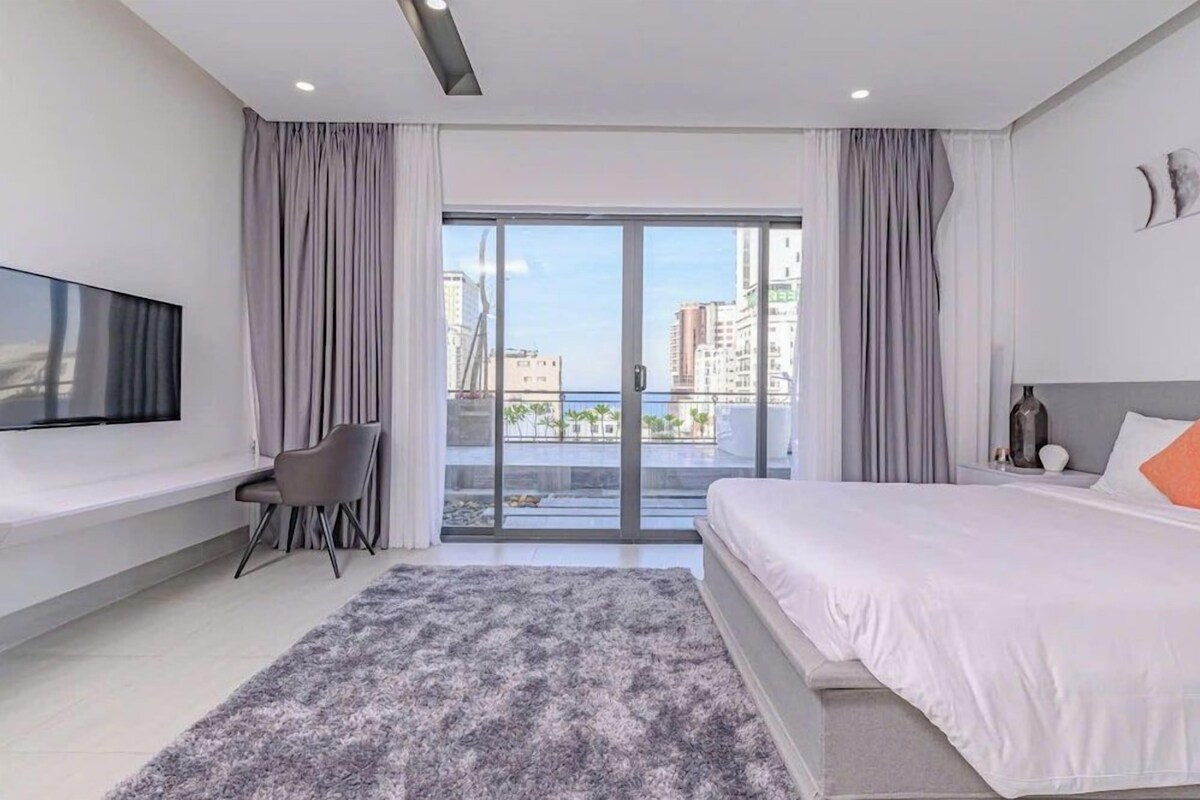 A modern hotel room with a large bed, a flat-screen TV, a desk, and a view of the city through large windows.