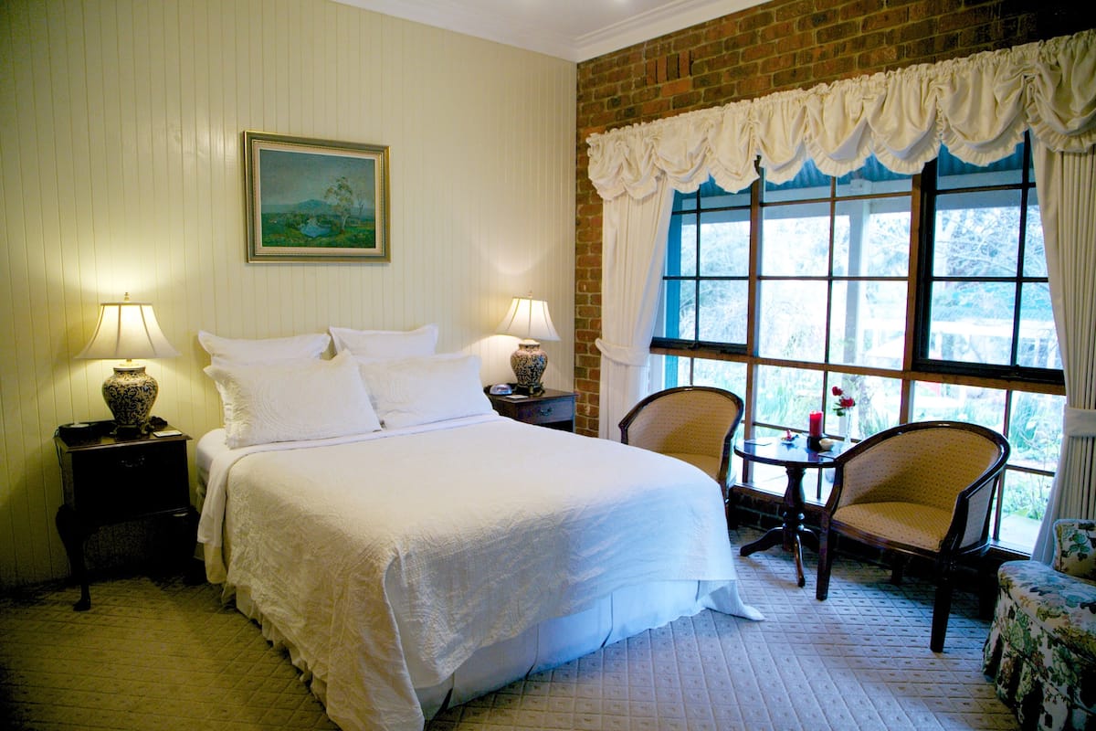 A neatly made bed with white linens, two bedside tables with lamps, a chair, and a window with curtains.