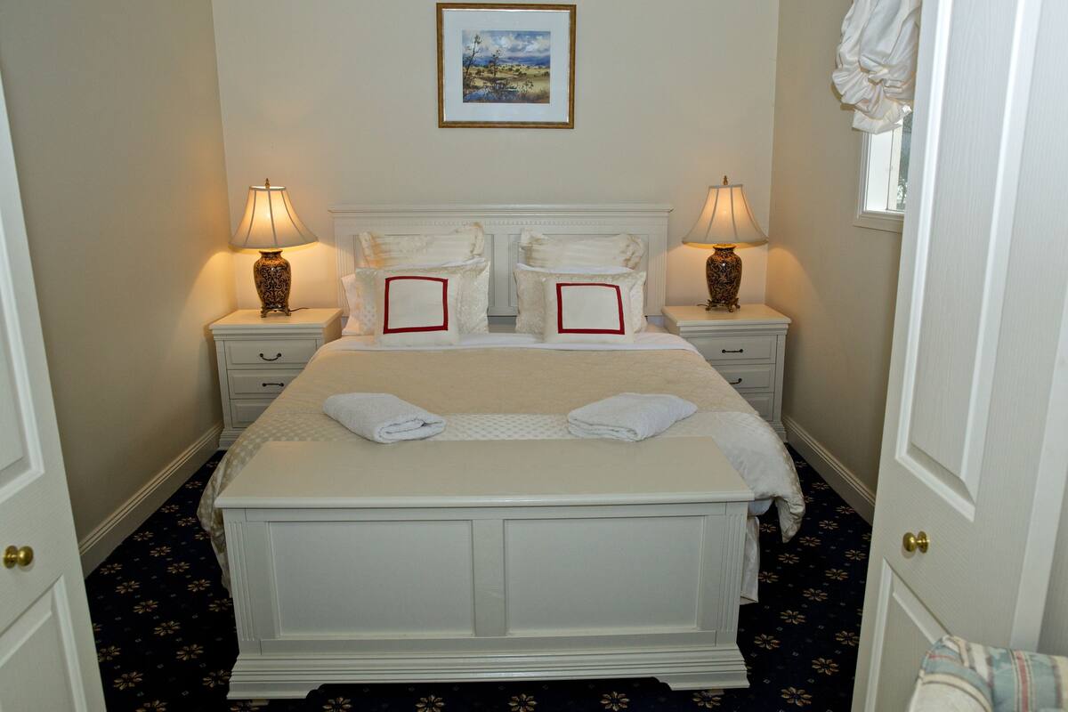 A neatly made bed with white linens and two pillows, flanked by bedside tables with lamps, in a room with a dark patterned carpet and a framed picture on the wall.