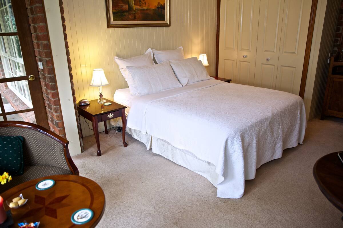 A neatly made bed with white linens, a bedside table with a lamp, and a chair with a floral cushion.