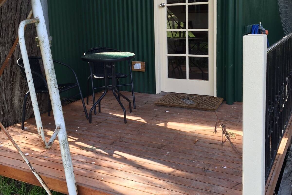 A wooden deck with a table and chairs, a door with a window, and a green building with a white door.