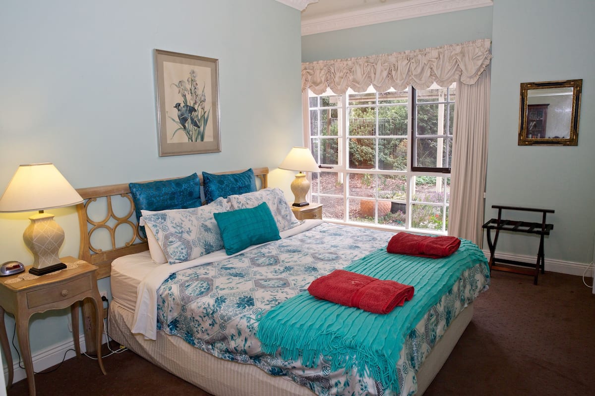 A bedroom with a large bed, two bedside tables with lamps, a window with curtains, a framed picture on the wall, and a small table with a lamp.