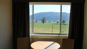 A room with a window overlooking a grassy field and mountains. There is a round wooden table with two chairs.