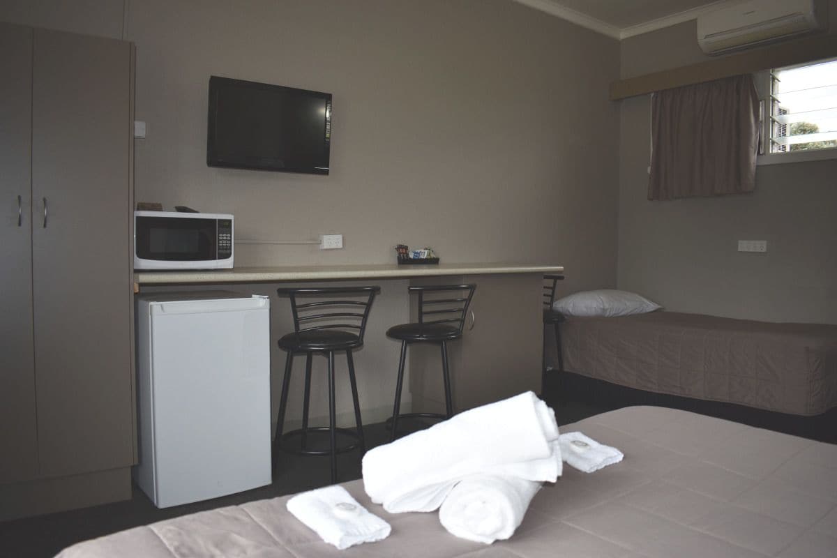 Habitación de hotel compacta con una cama, una pequeña kitchenette con microondas, refrigerador y televisor montado en la pared. Se aprecia una ventana con cortinas y una sola silla cerca de la kitchenette.