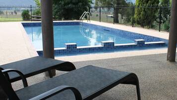 Una piscina con borde de azulejos, cercada por una valla metálica, y un área de descanso cubierta con bancos.