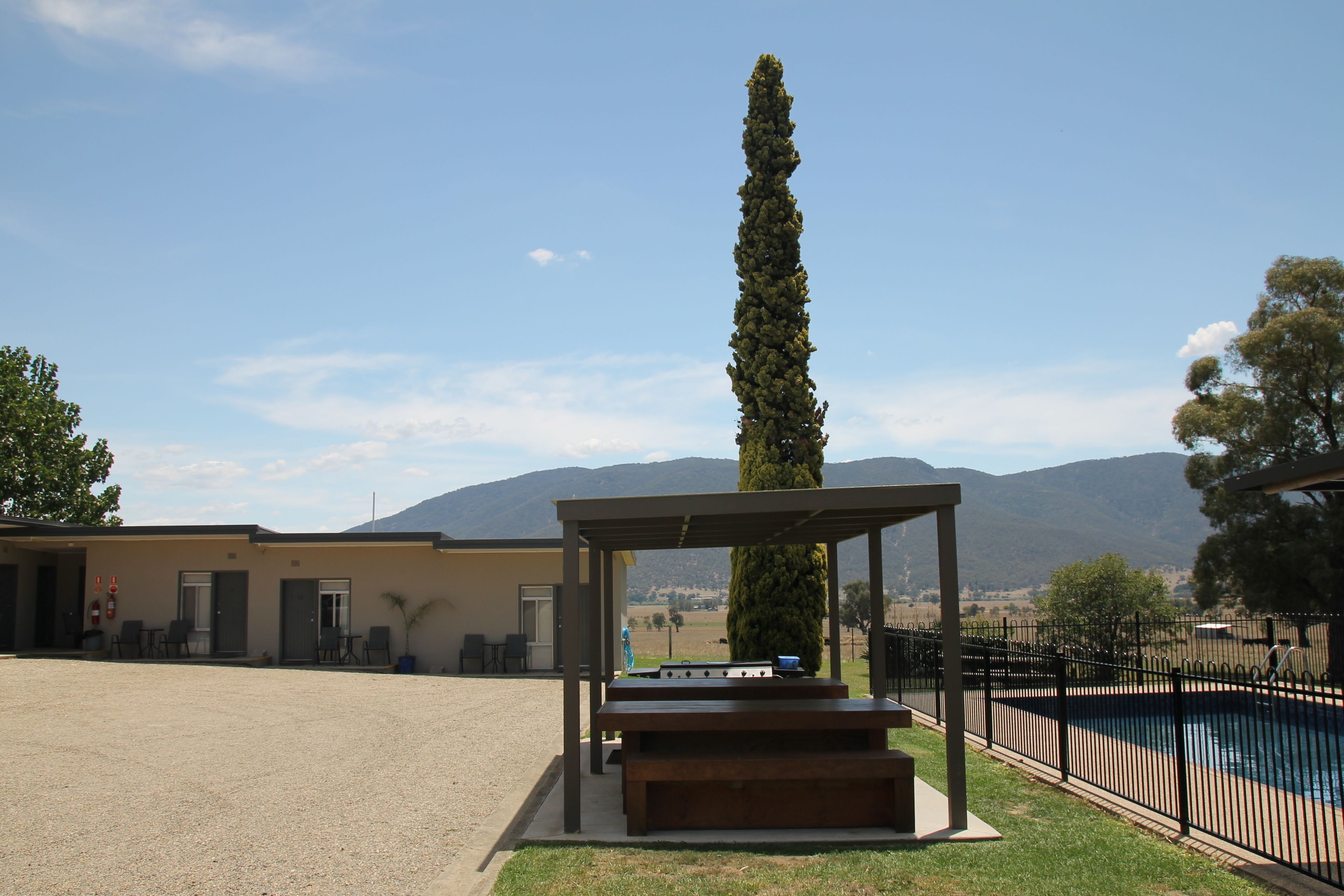 Uma área de estar coberta ao ar livre com churrasqueira, uma árvore alta e uma piscina com cerca.