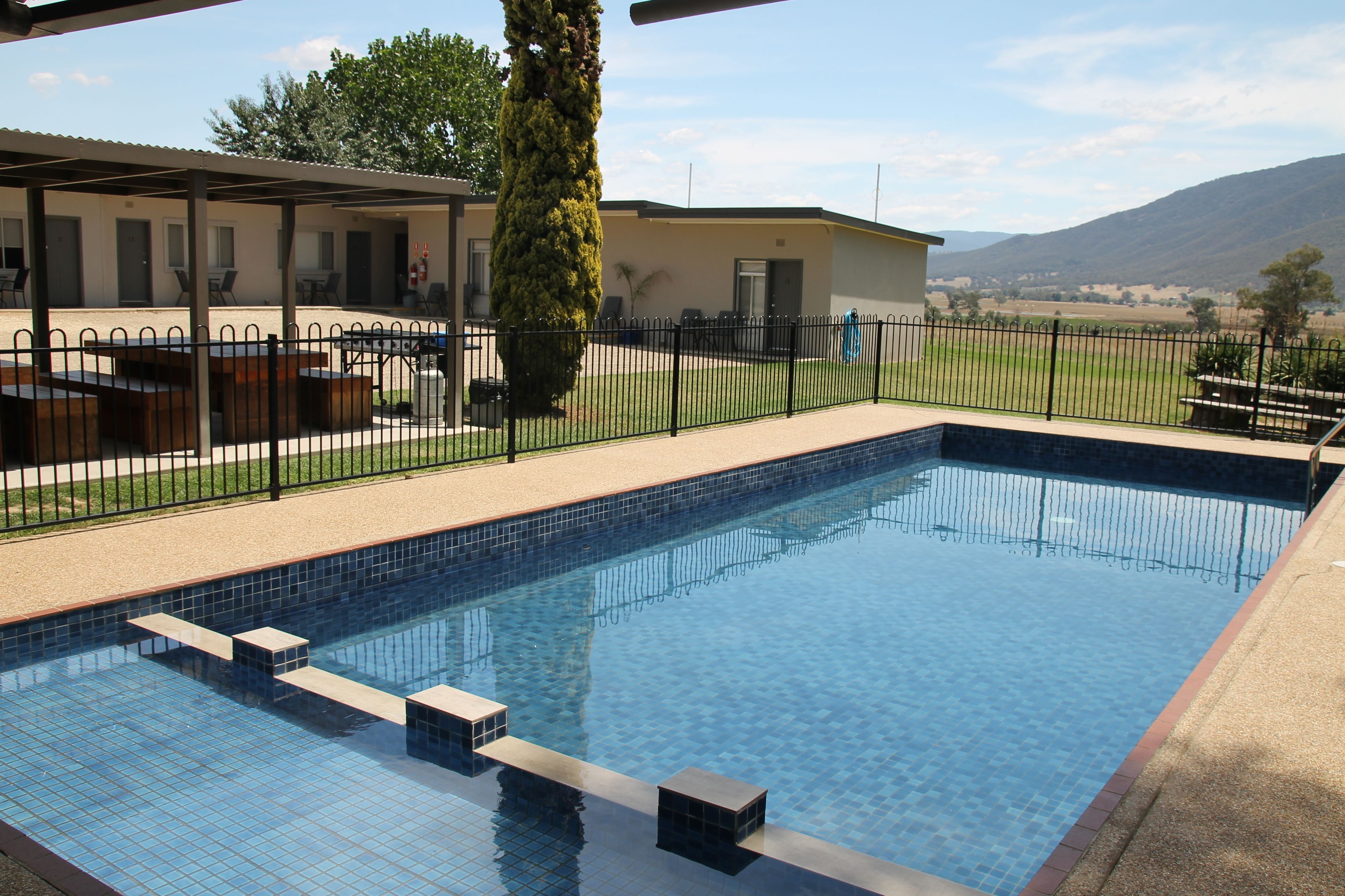 Una piscina con escalones de acceso, rodeada por una valla y un edificio con mobiliario exterior.