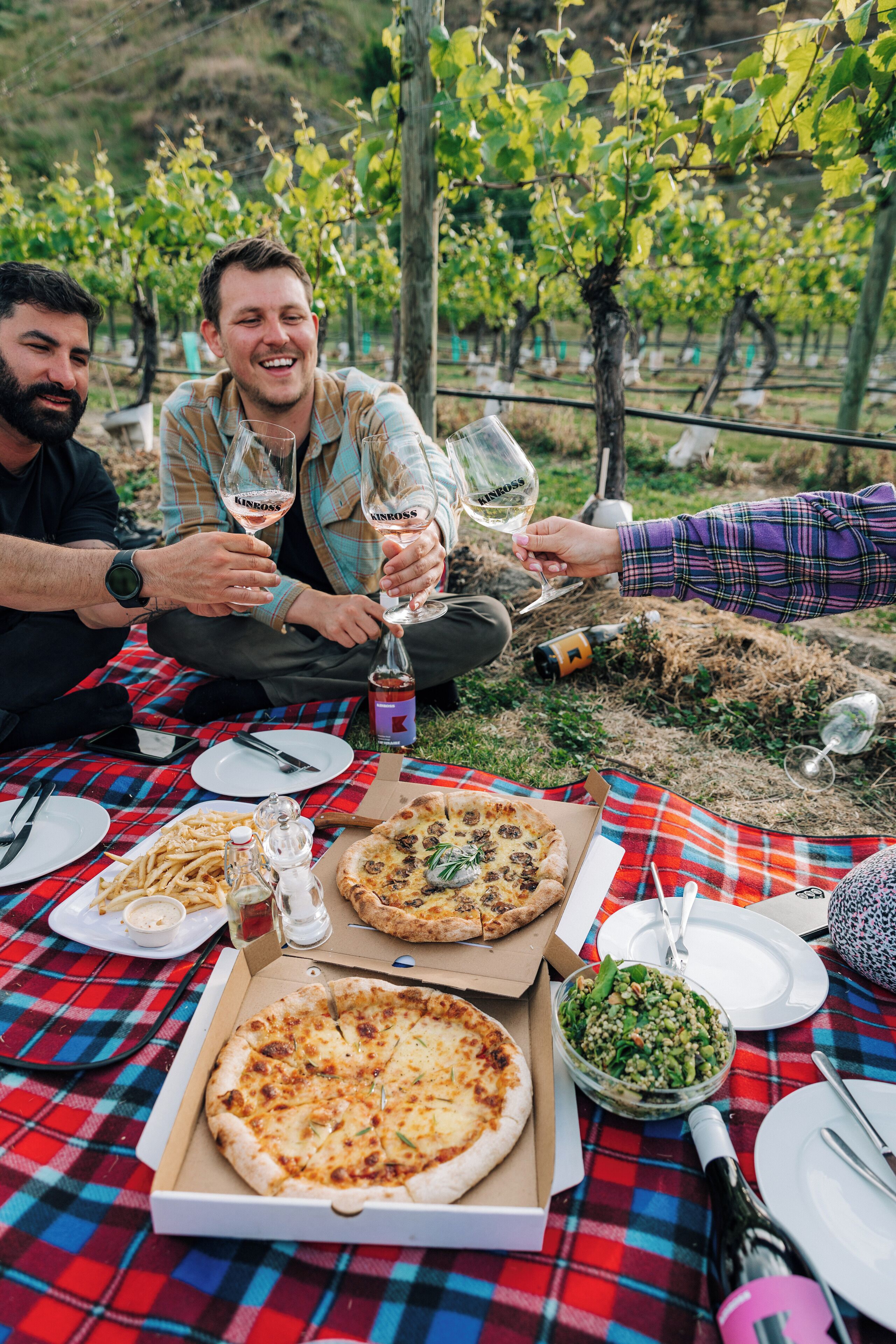 A group of people enjoying a meal outdoors with pizza and wine, seated on a checkered tablecloth.