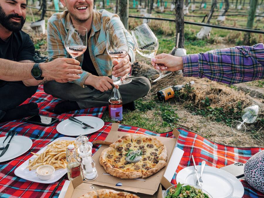Un groupe de personnes partage un repas en plein air, composé de pizzas et de vin, et sont assis autour d’une table recouverte d’une nappe à carreaux.