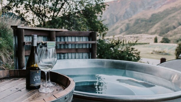 Een houten vat met een fles wijn en een glas op een houten tafel, naast een hottub met uitzicht op een berg in de verte.