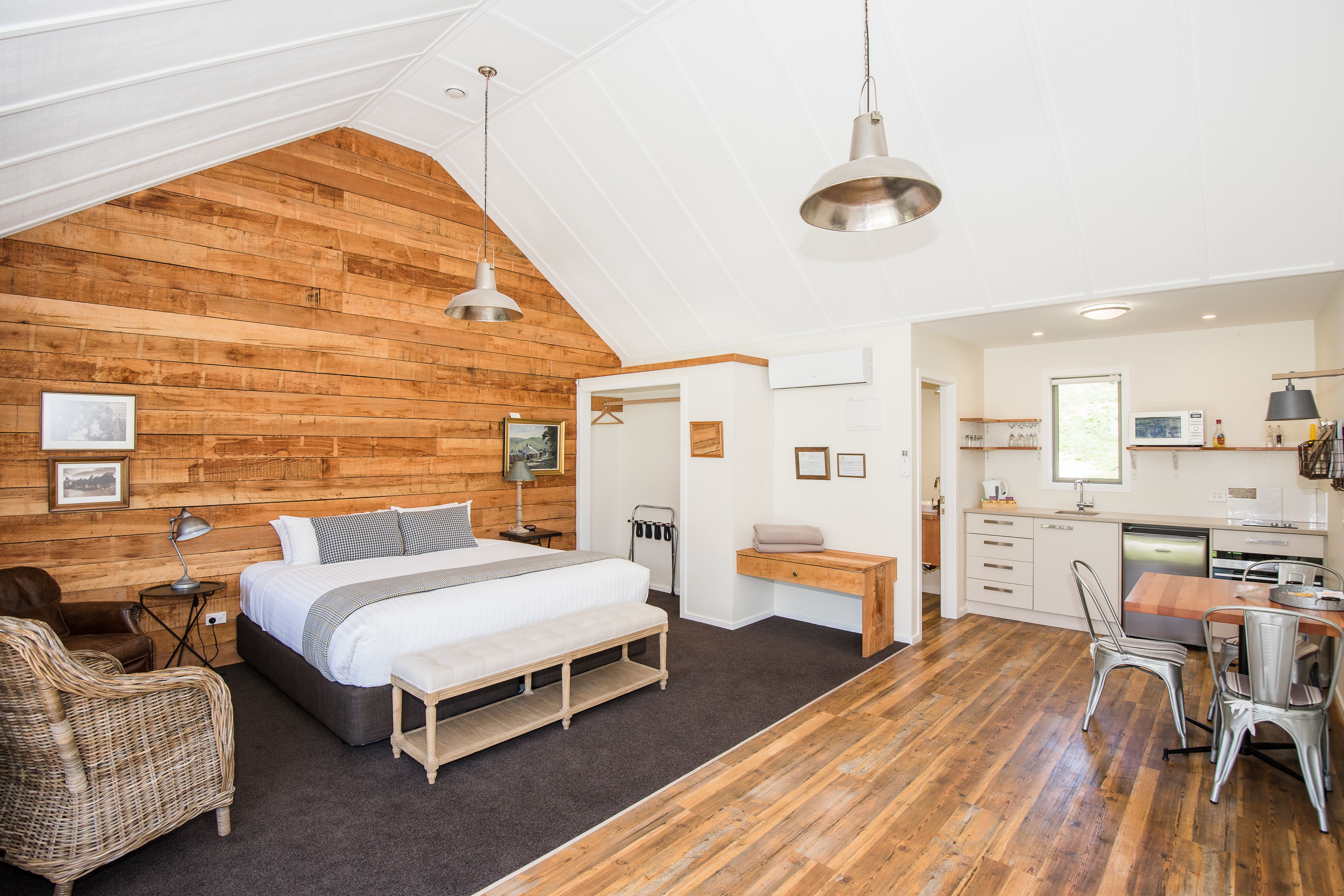 A bedroom with a large bed, a wooden ceiling, a desk, and a kitchenette.