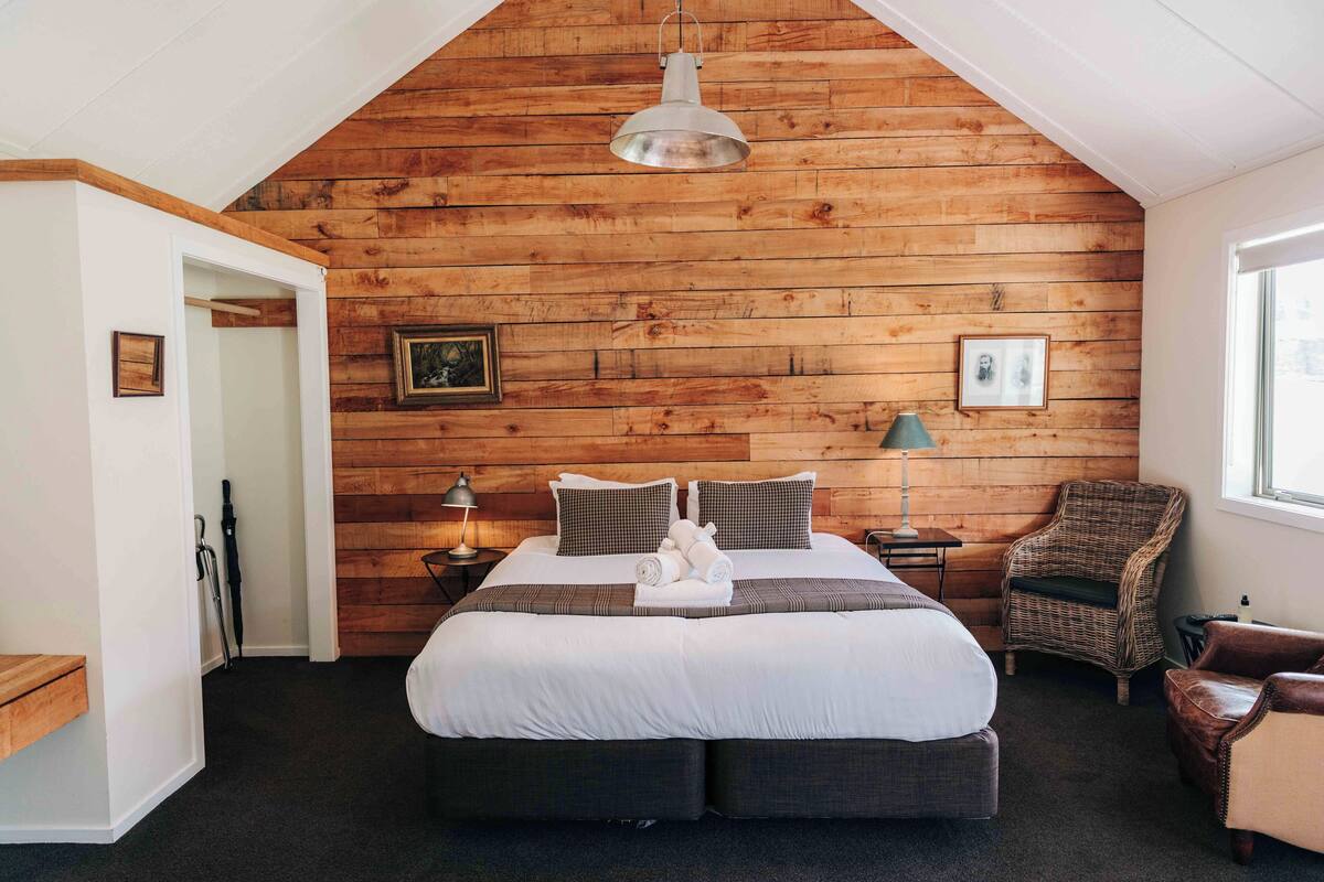 Un dormitorio con techo de madera inclinado, una cama grande con almohadas, una silla y una lámpara.
