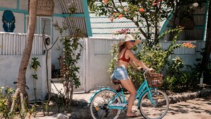 A woman riding a turquoise bicycle with a basket, parked in a courtyard with a white building, palm trees, and flowering plants. - 7SEAS Cottages (Gili Air)