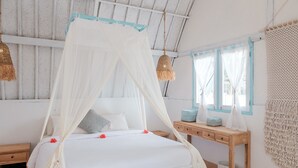 A bedroom with a canopy bed, wooden nightstand, bedside table with decorative items, and a view of the outside through a window with curtains. - 7SEAS Cottages (Gili Air)