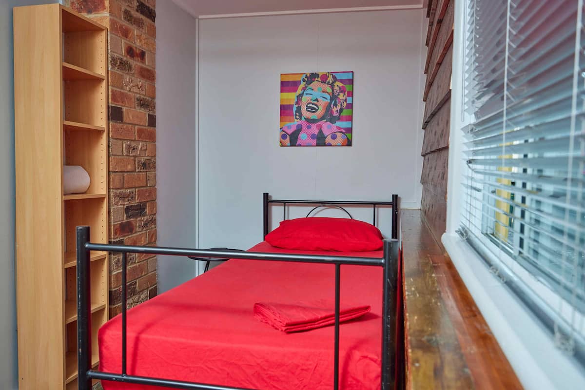 A single bed with a red mattress and pillow, a wooden shelf unit, a brick wall, and a window with blinds.