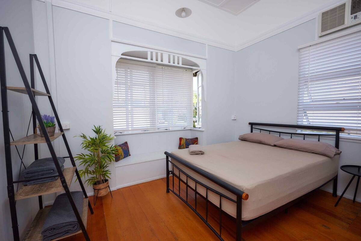 A bedroom with a bed, a window with blinds, a shelf with folded towels, a potted plant, and a ladder.