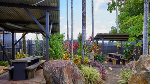 A covered outdoor area with stone benches, a wooden picnic table, and a variety of tropical plants. - Cairns City Backpackers Hostel (Parramatta Park)