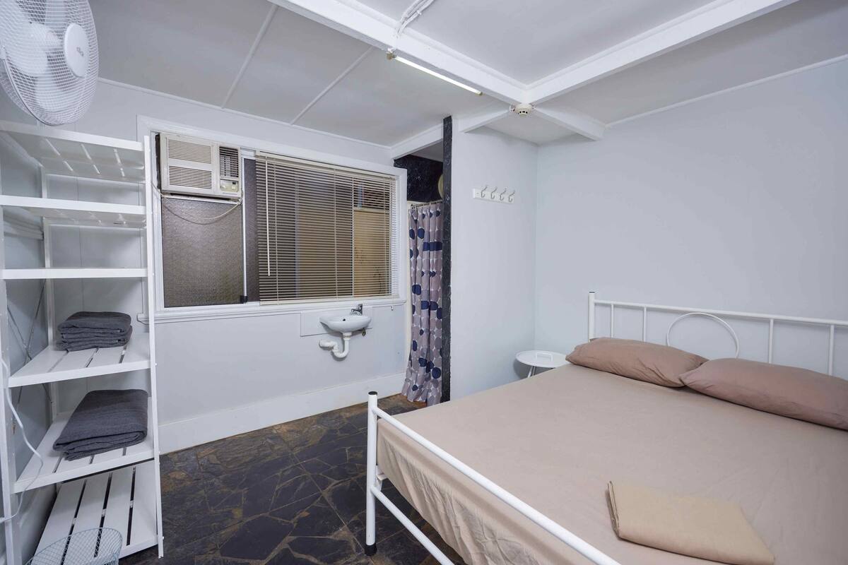 A bedroom with a bed, a white fan, a window with blinds, a sink, and a storage unit with shelves.