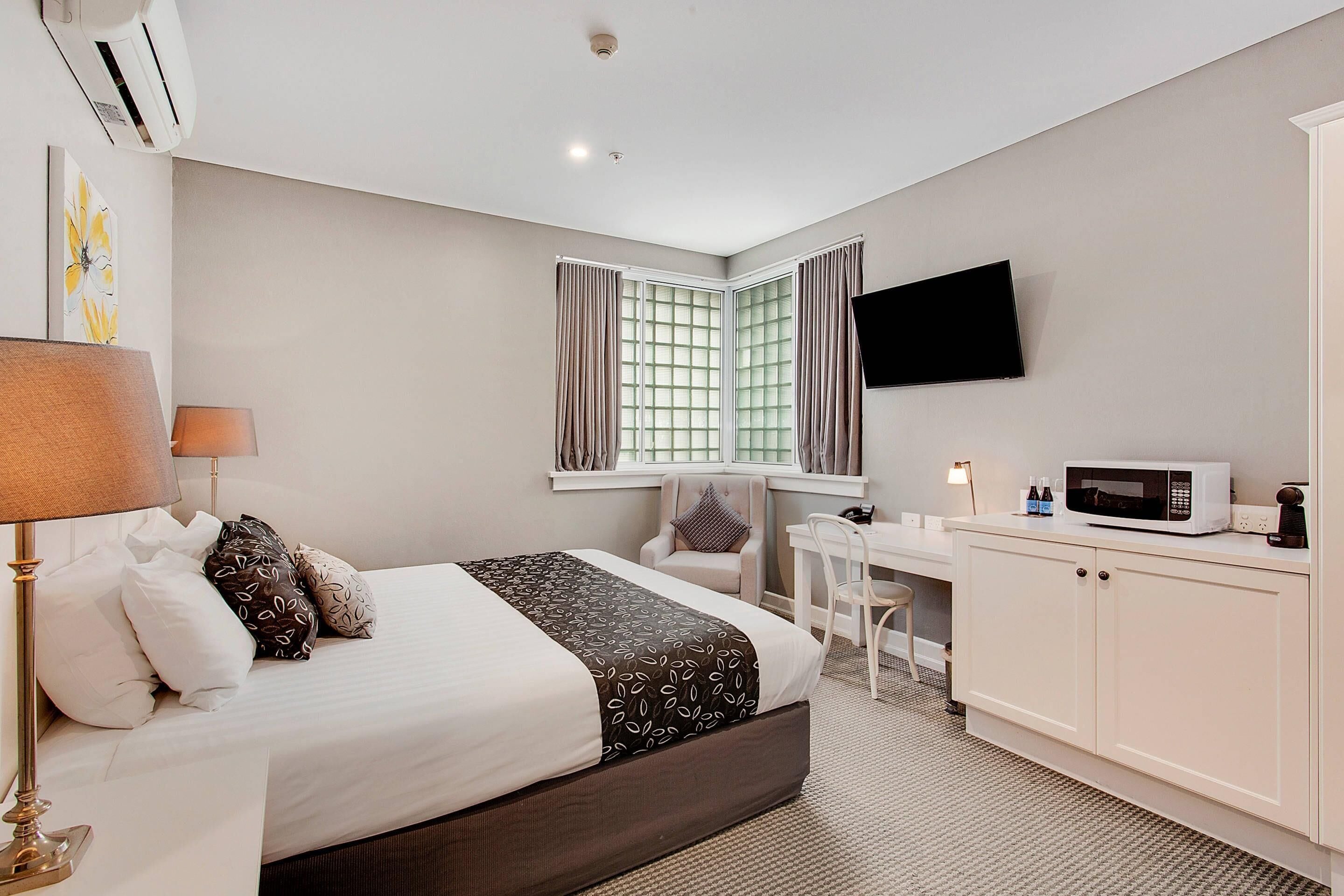 Ein Hotelzimmer mit Bett, Nachttisch, Lampe, Fernseher, Mikrowelle und Schreibtisch mit Stuhl.