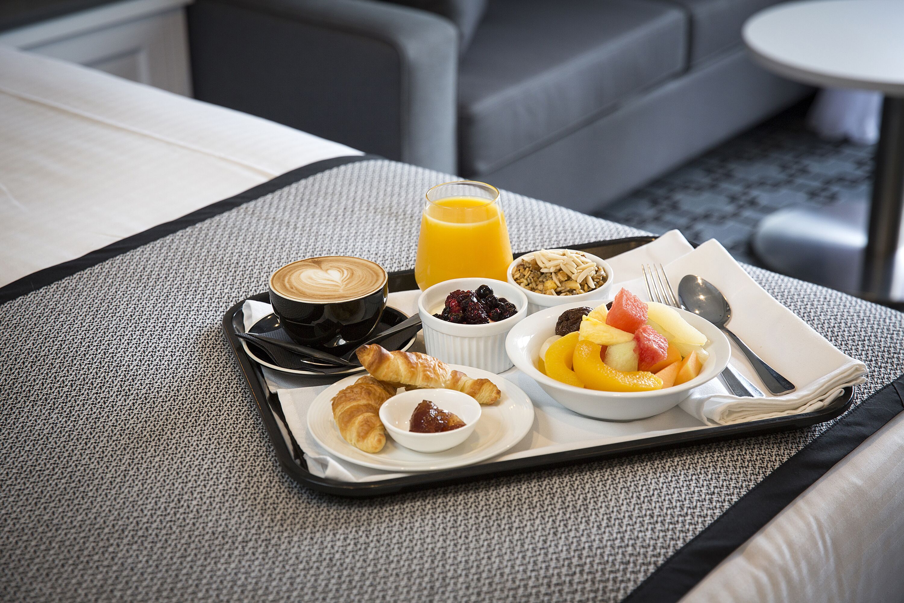 A breakfast tray with croissants, fruit, granola, a glass of orange juice, and a cup of coffee on a textured bedspread.