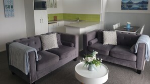 A modern living room with two sofas, a coffee table with flowers, a kitchen area with a green backsplash, and a painting of a mountain landscape. - Little Haven (Taupo)