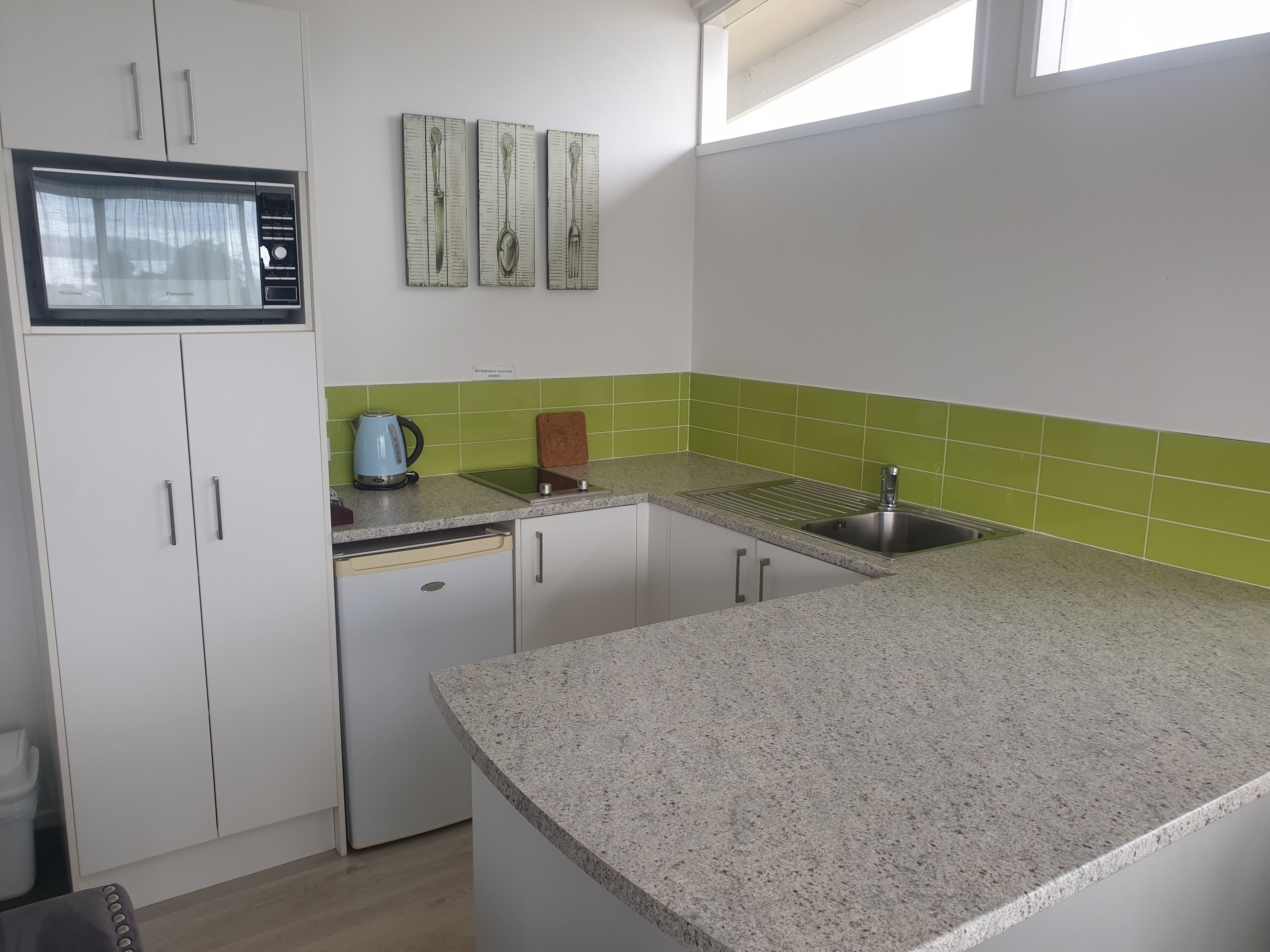 Une cuisine avec des meubles blancs, un four à micro-ondes, une bouilloire, un évier et des carreaux verts.