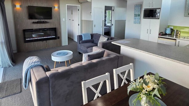 A modern living room with a sofa, a coffee table, and a TV mounted on a wall. There is a kitchen area with cabinets and a microwave. - Little Haven (Taupo)