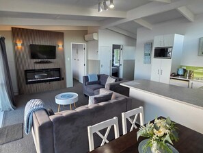 A modern living room with a sofa, a coffee table, and a TV mounted on a wall. There is a kitchen area with cabinets and a microwave. - Little Haven (Taupo)