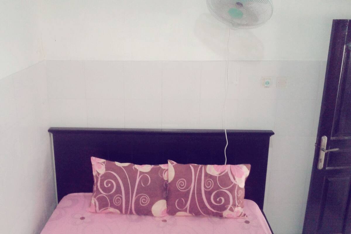 Une petite chambre avec un lit simple, dotée d’une tête de lit sombre, d’une couvre-lit rose et d’un coussin décoratif. Un ventilateur fixé au mur est visible au-dessus du lit.