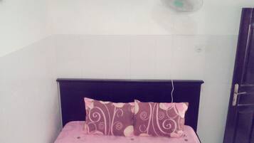 Una habitación pequeña con cama individual, cabecera oscura, colcha rosa y almohada decorativa. Se ve un ventilador de pared sobre la cama.