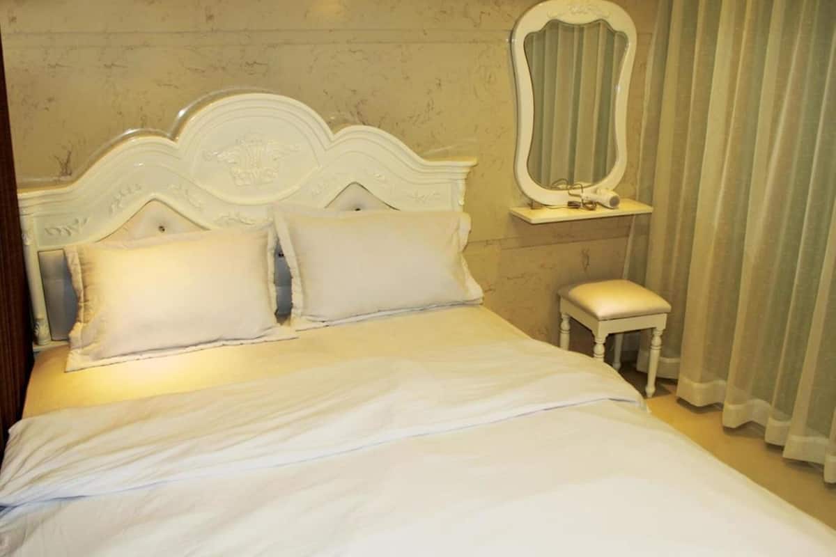 Uma cama com cabeceira branca e duas almofadas, um banquinho branco pequeno e um espelho sobre uma cômoda com pia.