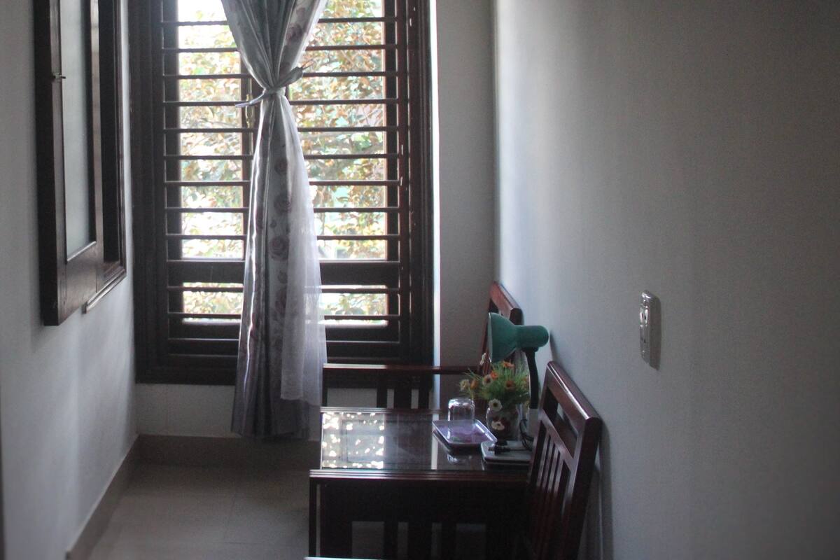 A room with a dining table, chairs, a hanging light fixture, a window with curtains, and a framed picture on the wall.
